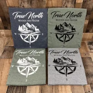 Four True North compass-themed t-shirts in different colors laid out on a wooden floor.