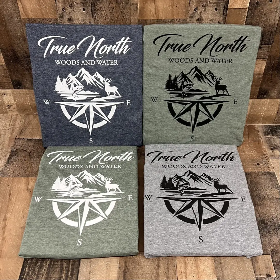 Four True North compass-themed t-shirts in different colors laid out on a wooden floor.