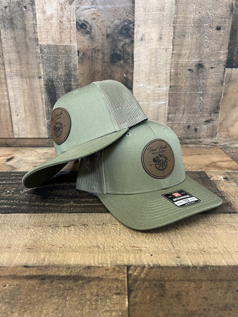 Two olive green baseball caps with round leather patches on the front.
