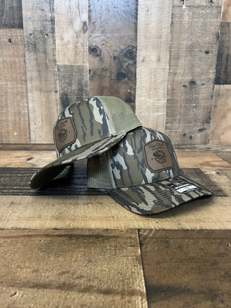 Two camouflage-patterned baseball caps with leather patches on a wooden surface.