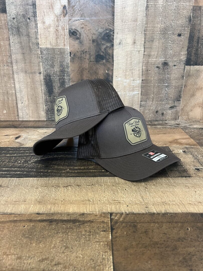 Two gray baseball caps with embroidered patches on a wooden surface.