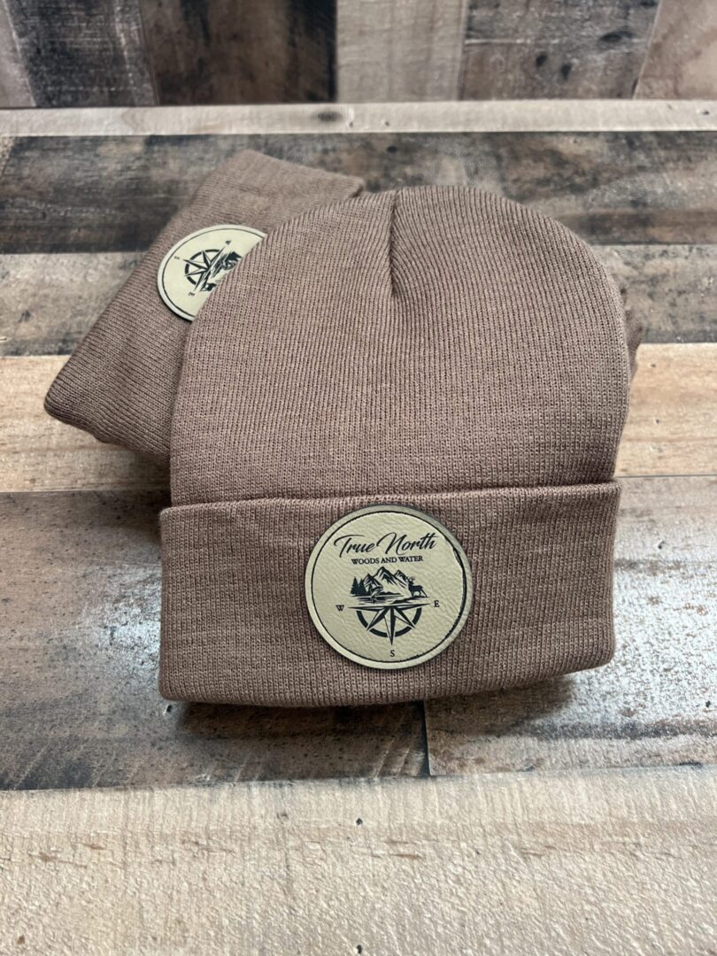 Two beige knit beanies with a round patch on the cuff.